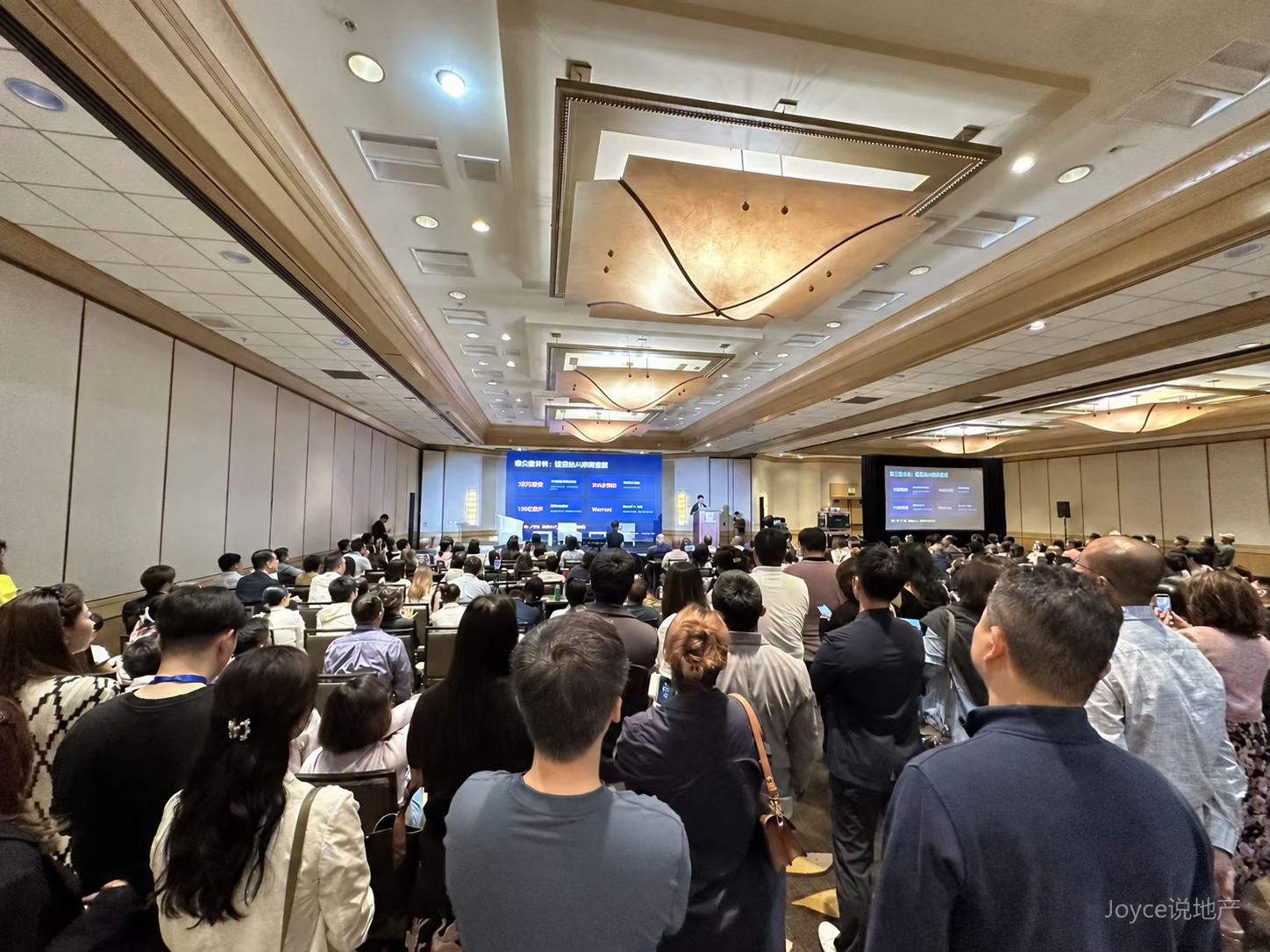 李尚龙在AI资金流向主题演讲现场，右下角带“Joyce说地产”暗标 / Li Shanglong speaking at an AI capital-flows event, with a subtle Joyce Says Real Estate watermark